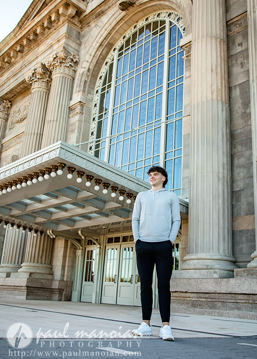 A young man in a light hoodie and black pants stands smiling in front of a large historic building with tall arched windows and columns, captured during a Detroit senior pictures session. The photo is credited to Paul Manoian Photography.