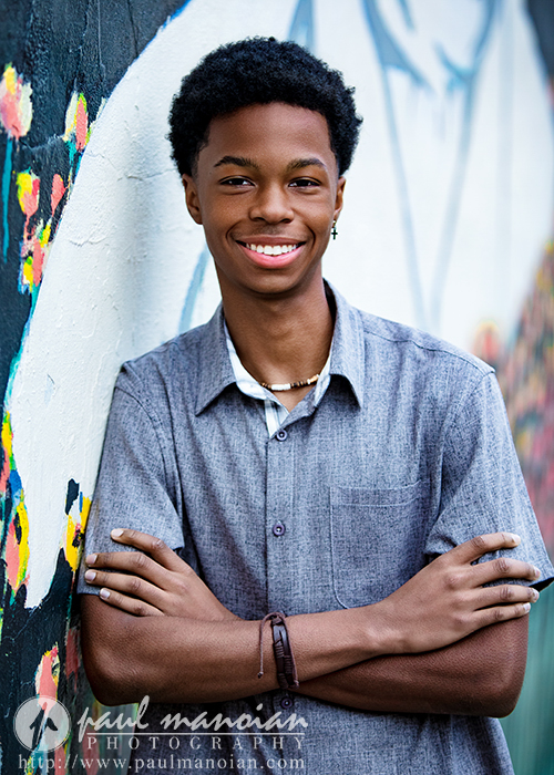 A smiling high school senior with short curly hair stands with arms crossed in front of a colorful mural during his Novi senior pictures session, wearing a gray button-up shirt, beaded necklace, and bracelet.