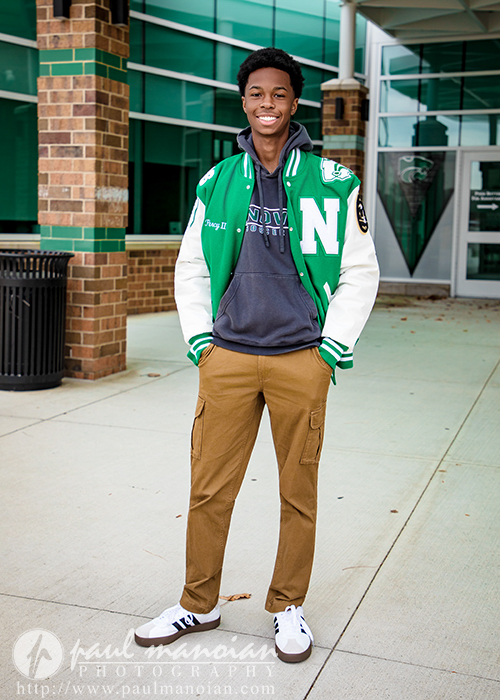 A smiling high school senior stands outside a school building during his Novi senior pictures session, wearing a green and white letterman jacket, dark hoodie, brown cargo pants, and white sneakers. He poses confidently with hands in his pockets.