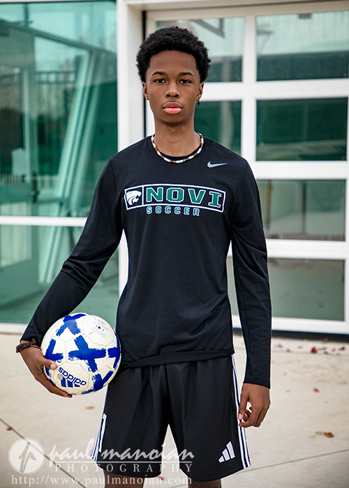 A young soccer player stands outside holding a blue and white soccer ball. He wears a black long-sleeve Novi Soccer shirt—perfect for his Novi senior pictures session—with black shorts and white stripes. A building with large glass windows is in the background.
