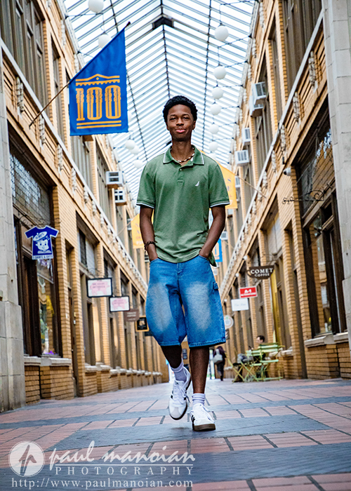 A high school senior wearing a green polo shirt and long denim shorts walks confidently through a glass-roofed urban corridor lined with yellow brick and shop signs during his Novi senior pictures session, hands in his pockets.