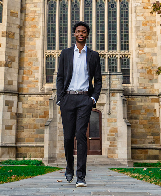 A young man in a suit walks confidently on a stone path in front of a large stone building with tall arched windows—an ideal scene for a Novi senior pictures session—surrounded by grass and trees.