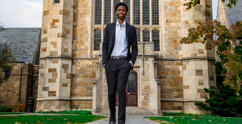 A young man in a suit walks confidently on a stone path in front of a large stone building with tall arched windows—an ideal scene for a Novi senior pictures session—surrounded by grass and trees.