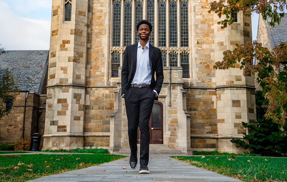 A young man in a suit walks confidently on a stone path in front of a large stone building with tall arched windows—an ideal scene for a Novi senior pictures session—surrounded by grass and trees.