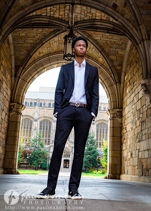 A high school senior in a black suit stands confidently under an arched stone walkway, capturing the timeless elegance of his Novi senior pictures session, with a historic building and greenery in the background.