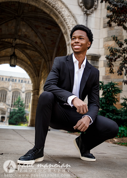 A high school senior in a black suit and sneakers smiles while crouching outdoors during his Novi senior pictures session, with an arched stone building and greenery in the background.