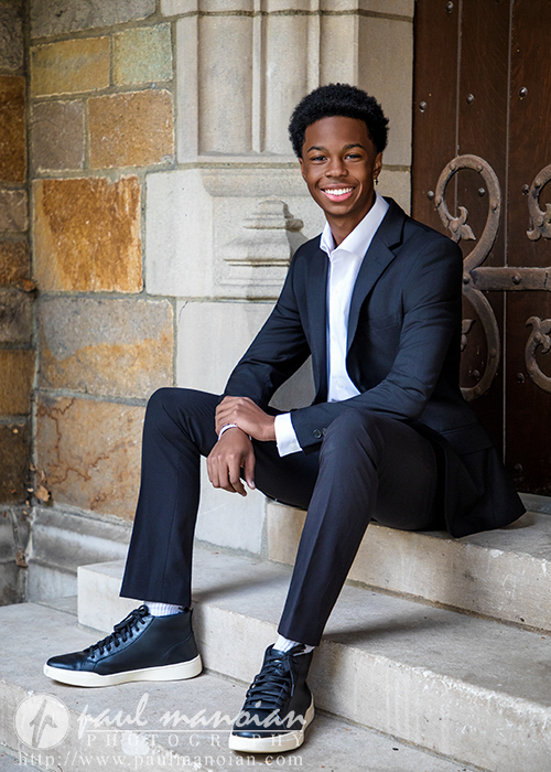 A high school senior wearing a black suit, white shirt, and black sneakers sits on stone steps in front of a wooden door, smiling at the camera during his Novi senior pictures session. The background features a stone wall with various shades of tan and gray.