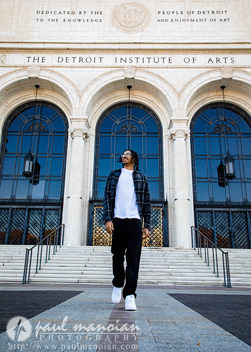 A high school senior in casual clothing walks down the steps in front of the Detroit Institute of Arts, grand arched entryways behind him—an iconic spot for a Detroit senior pictures session.