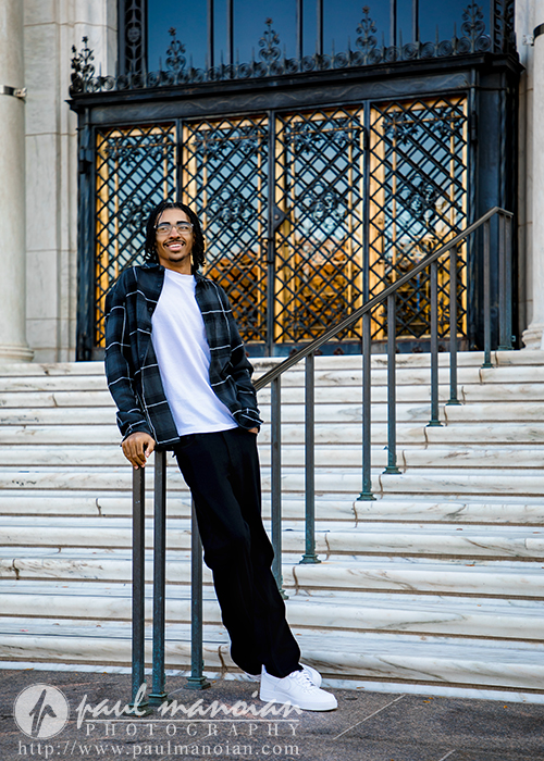 A high school senior with glasses, in a black plaid shirt over a white t-shirt, black pants, and white sneakers, leans casually on a railing before marble steps and ornate doors during his Skyline High School senior pictures session. Photography watermark is present.