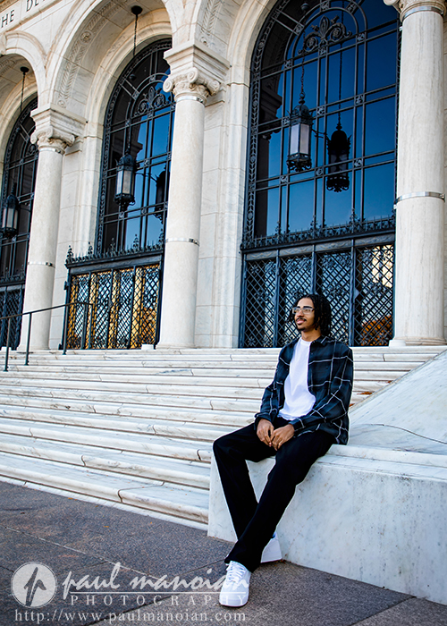 A high school senior in a plaid shirt sits on marble steps in front of a grand building with arched windows and iron gates, enjoying bright natural light during his Detroit senior pictures session.