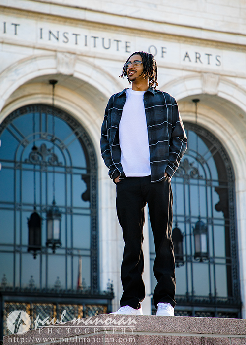 A high school senior wearing a plaid shirt, white t-shirt, and dark pants stands on steps in front of a building labeled "INSTITUTE OF ARTS," smiling and looking to the side during his Detroit senior pictures session. The building has large arched windows.