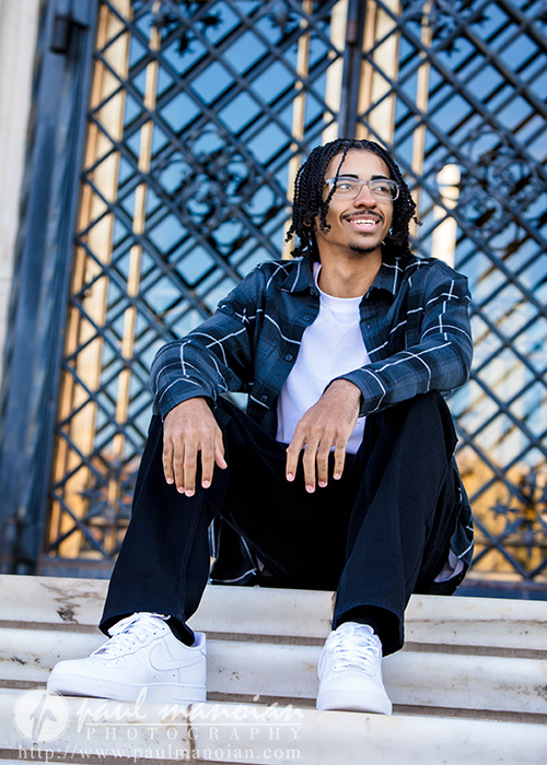 A high school senior with glasses and braided hair, wearing a plaid shirt over a white t-shirt, black pants, and white sneakers, sits on outdoor steps, smiling during his Detroit senior pictures session with a decorative iron gate in the background.