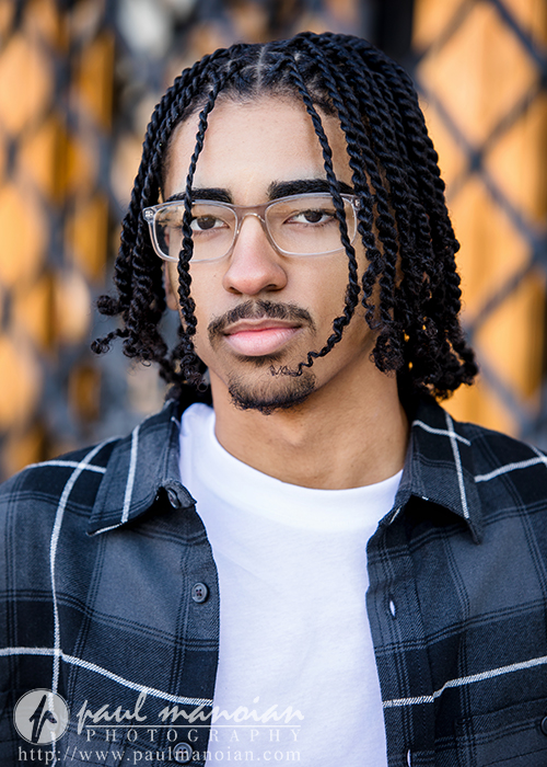 A high school senior with medium-length twisted hair, glasses, and a mustache, wearing a black and white plaid shirt over a white t-shirt, stands in front of a blurred outdoor background during his Detroit senior pictures session.