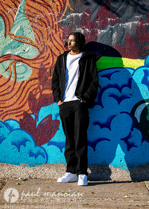 A high school senior in a black hoodie, black pants, and white sneakers stands against a colorful mural with abstract shapes and a large sun—perfect inspiration for a Detroit senior pictures session. He looks to the side with hands in his pockets, partially in shadow.