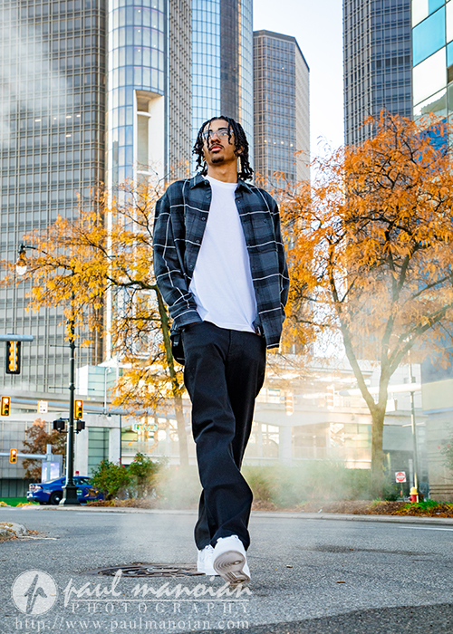 A high school senior in casual clothing walks confidently on a city street during his Detroit senior pictures session, with tall buildings and autumn trees in the background, steam rising from the ground.