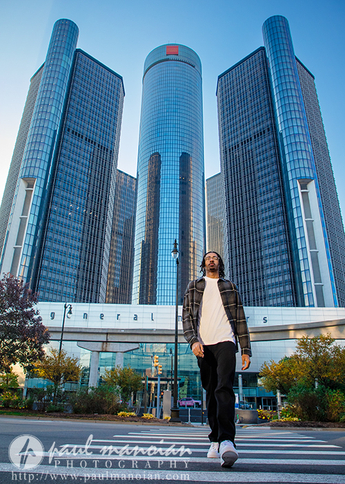 A high school senior in casual clothes walks across a crosswalk in front of the glass towers of the GM Renaissance Center in Detroit on a sunny day—an ideal spot for a Detroit senior pictures session framed by trees and greenery.