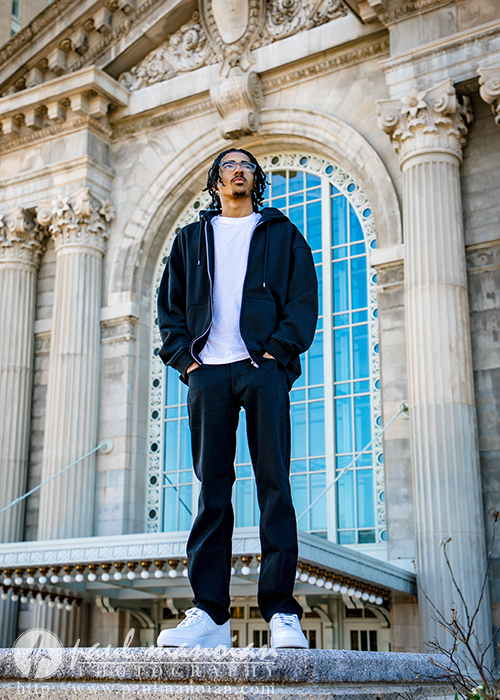 A high school senior in a black hoodie, white t-shirt, and black pants stands confidently in front of a historic building with large columns and an arched window during his Detroit senior pictures session.