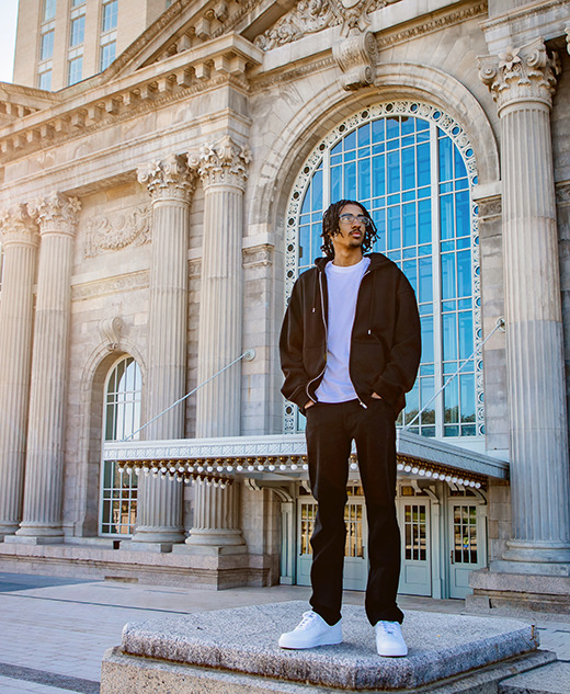 A man in casual clothing stands on a stone pedestal in front of a grand building with tall columns and large arched windows, sunlight shining from the left during a Detroit senior pictures session.