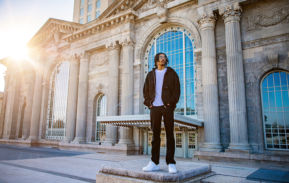 A man in casual clothing stands on a stone pedestal in front of a grand building with tall columns and large arched windows, sunlight shining from the left during a Detroit senior pictures session.