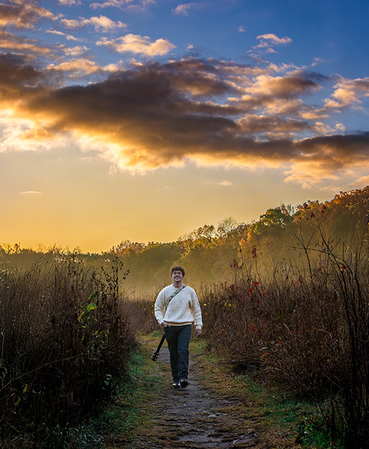 A person walks along a narrow path through tall grass and shrubs at sunrise or sunset, with a dramatic cloudy sky and warm golden light in the background—perfect for a South Lyon senior pictures session.