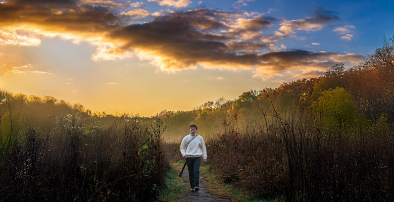 A person walks along a narrow path through tall grass and shrubs at sunrise or sunset, with a dramatic cloudy sky and warm golden light in the background—perfect for a South Lyon senior pictures session.