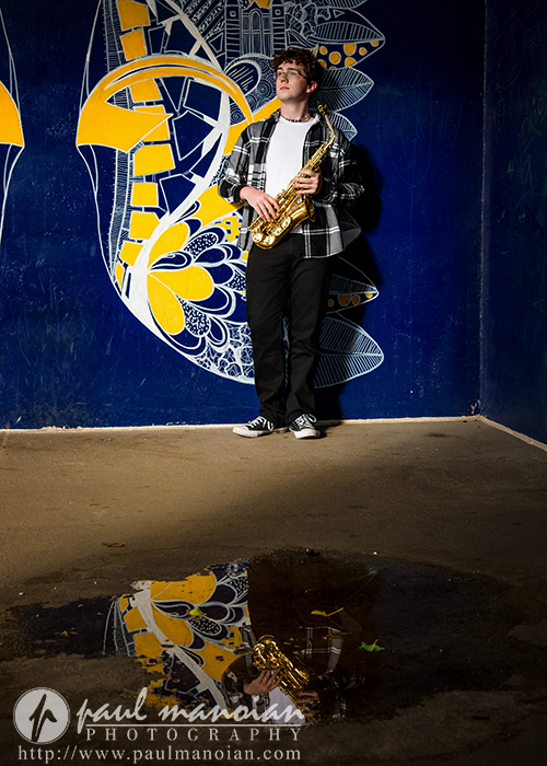 A young person holding a saxophone stands against a blue mural with yellow and white abstract patterns, captured during a South Lyon senior pictures session. The instrument and mural are beautifully reflected in a puddle on the ground.