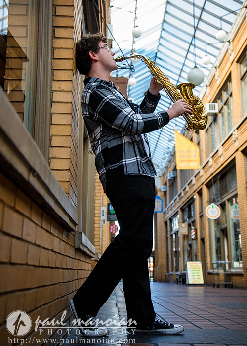 A young man wearing a plaid shirt and black pants plays a saxophone while leaning against a brick wall in an indoor shopping arcade with a glass ceiling during his South Lyon senior pictures session.