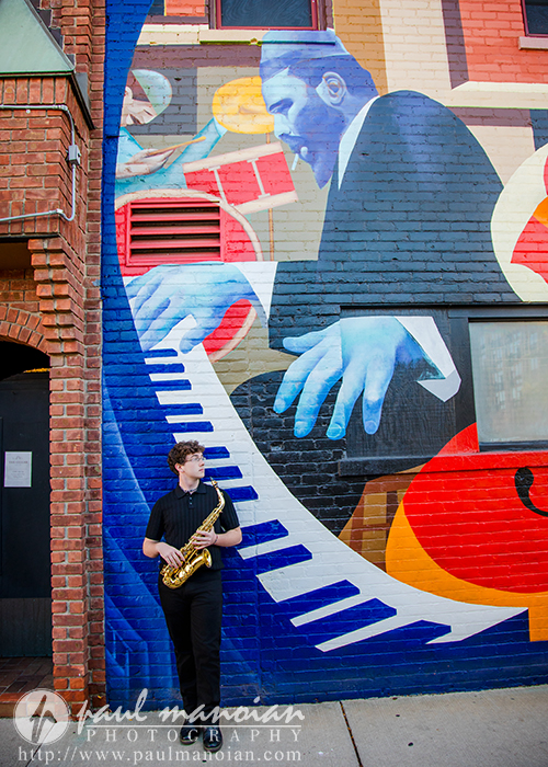 A person holding a saxophone stands against a brick wall featuring a colorful mural of a man playing piano keys, making this the perfect spot for a South Lyon senior pictures session with vibrant blue, red, yellow, and white accents.