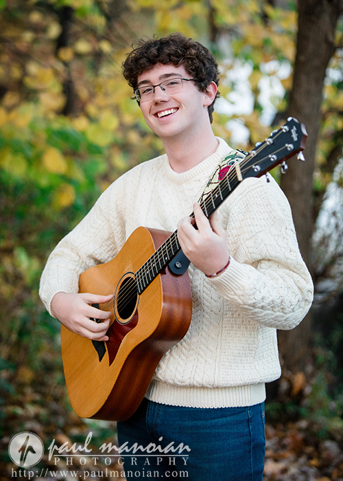 A young person with curly hair, glasses, and a cream sweater is smiling while playing an acoustic guitar outdoors during a South Lyon senior pictures session, surrounded by autumn trees with yellow leaves.
