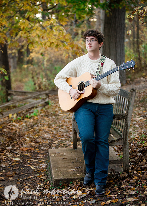 A young man with curly hair, wearing glasses, a cream sweater, and jeans, stands outdoors in a wooded area during his South Lyon senior pictures session, playing an acoustic guitar next to a wooden bench on a path covered with fallen leaves.