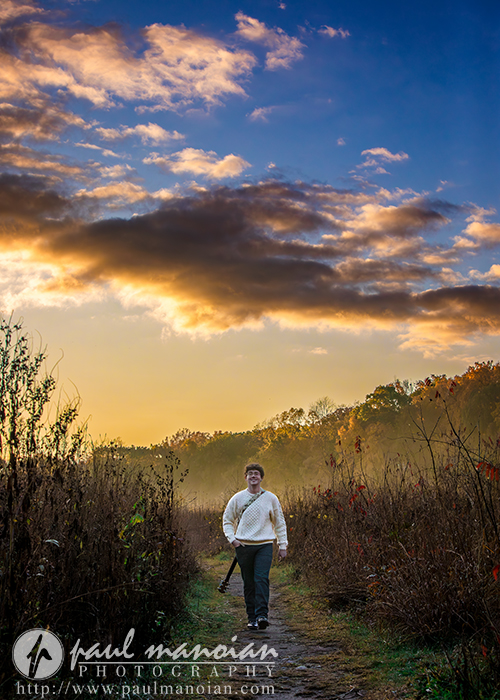 A person in a white sweater walks along a dirt path through tall grass at sunrise or sunset, dramatic clouds and golden light overhead—perfect inspiration for a South Lyon senior pictures session. Trees line the horizon in the background.