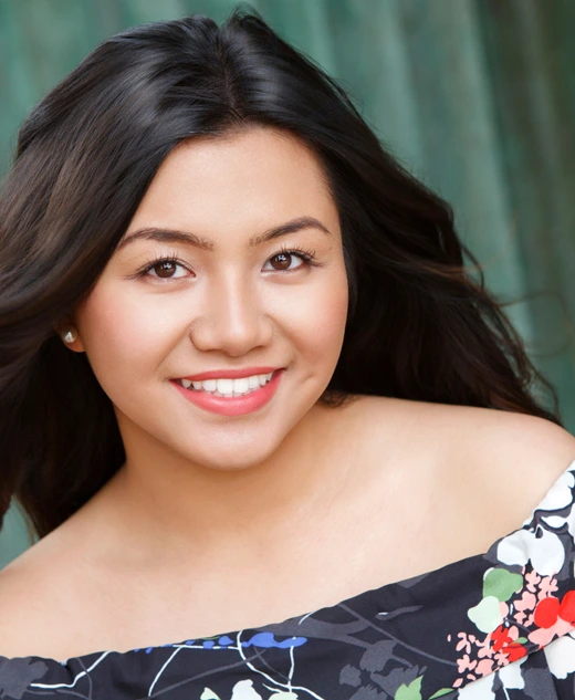 A high school senior girl with long dark hair smiles at the camera. She is wearing an off-the-shoulder floral dress and stands in front of a teal-colored textured background.