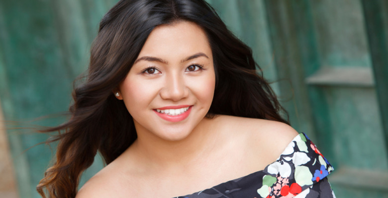 A high school senior girl with long dark hair smiles at the camera. She is wearing an off-the-shoulder floral dress and stands in front of a teal-colored textured background.
