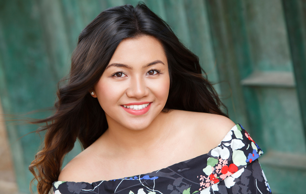 A high school senior girl with long dark hair smiles at the camera. She is wearing an off-the-shoulder floral dress and stands in front of a teal-colored textured background.