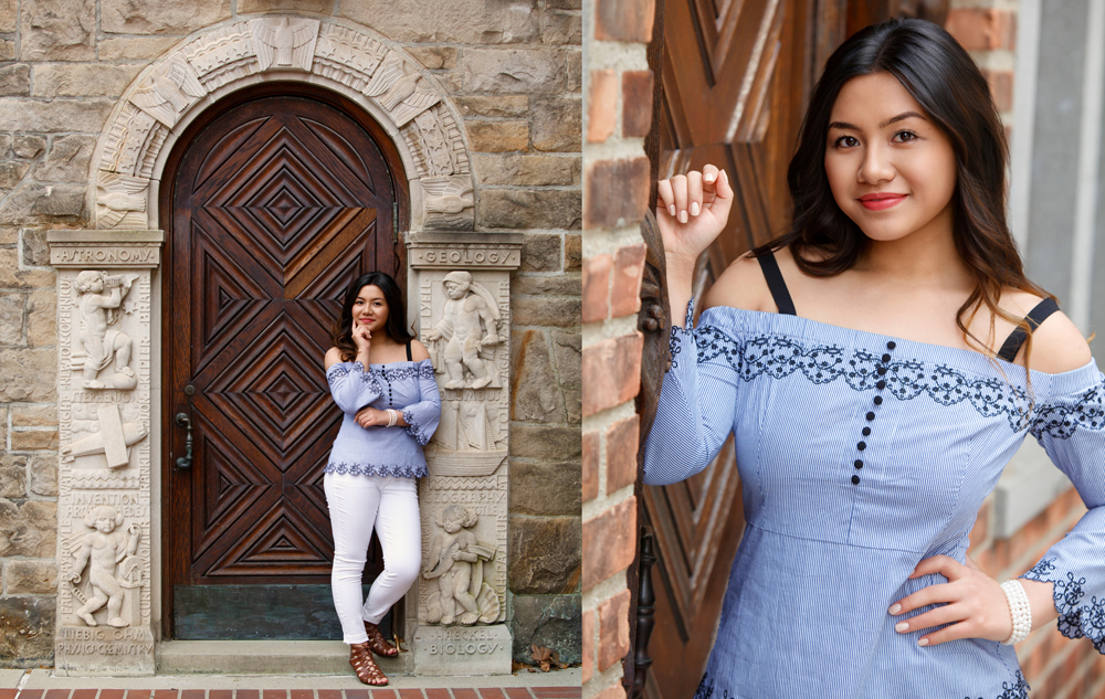 A young woman wearing a light blue off-shoulder top and white pants poses confidently in front of a decorative stone doorway and beside a brick wall, smiling at the camera.