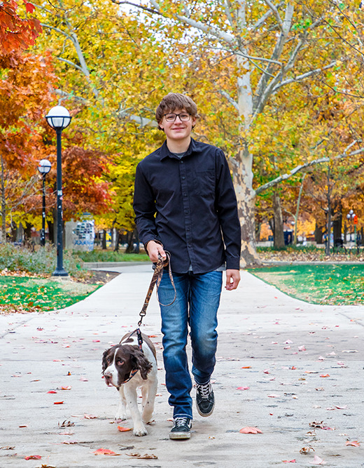 A young man wearing glasses, a black shirt, and jeans walks a brown and white dog on a leash along a tree-lined path covered with autumn leaves. The trees have orange, yellow, and green foliage.
