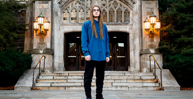 A person with long hair, wearing glasses, a blue shirt, and black pants stands in front of a grand stone building during their Livonia senior pictures session, framed by arched doorways and ornate lanterns.