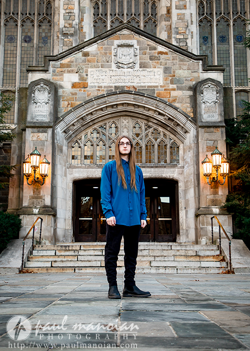 A person with long hair, wearing a blue shirt and black pants, stands on stone steps in front of a large, ornate building with gothic architecture and arched doorways—an elegant backdrop for a Livonia senior pictures session. Two lit lanterns flank the entrance.