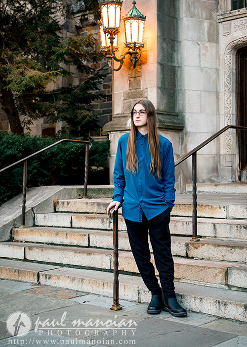 A person with long hair, glasses, and a blue shirt stands on stone steps outside a historic building during their Livonia senior pictures session, holding a railing. A lit lamp and carved stone details are visible in the background.