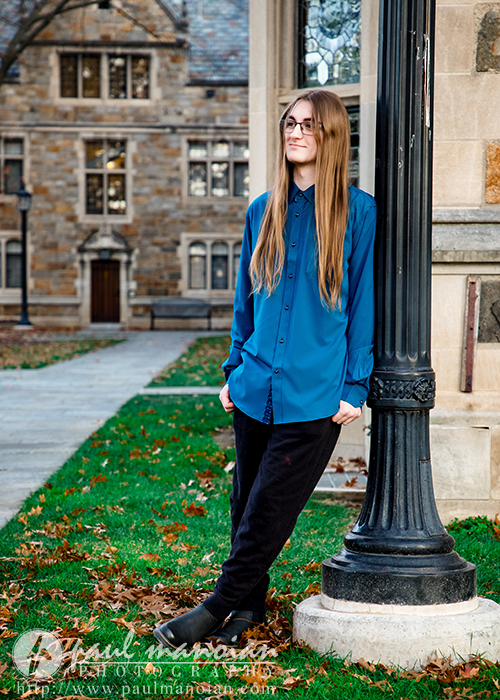 A person with long hair, glasses, and a blue shirt leans against a black lamppost outside a stone building with arched windows during a Livonia senior pictures session. Fallen leaves and a green lawn are visible in the background.