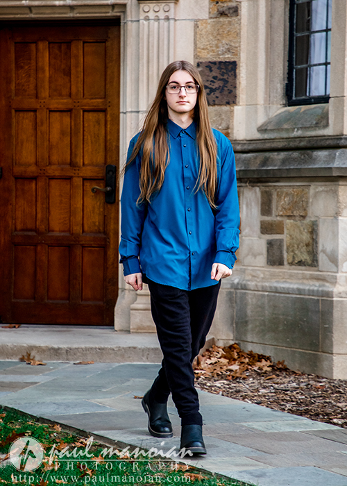 A person with long brown hair, glasses, and wearing a blue button-up shirt and black pants walks on a sidewalk near a stone building with a wooden door during a Livonia senior pictures session.