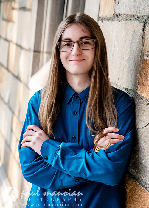 A person with long, straight hair, glasses, and a blue button-up shirt stands with arms crossed, smiling slightly against a stone wall during a Livonia senior pictures session.
