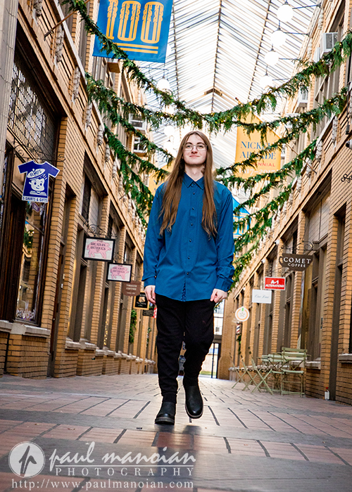 A person with long hair, glasses, a blue shirt, and black pants walks down a covered brick alleyway decorated with hanging greenery and signs—an inviting scene perfect for a Livonia senior pictures session, with bright lighting and cozy tables along the sides.