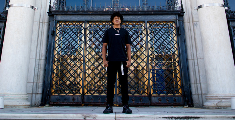 A young person in black clothes and a hat stands on marble steps before ornate metal gates at a grand building, capturing a striking moment for their Macomb senior pictures session.