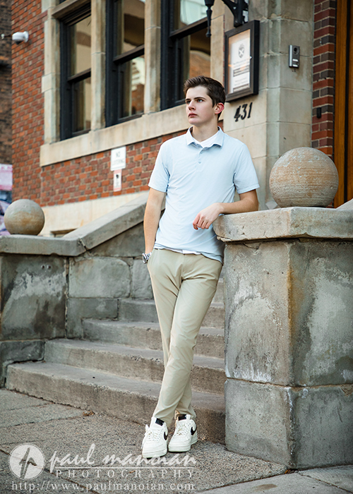A young man in a light blue polo and beige pants stands casually, leaning on a stone railing by outdoor steps in front of a brick building—an ideal pose for an Oxford senior pictures session. He looks to the side, with one hand in his pocket.