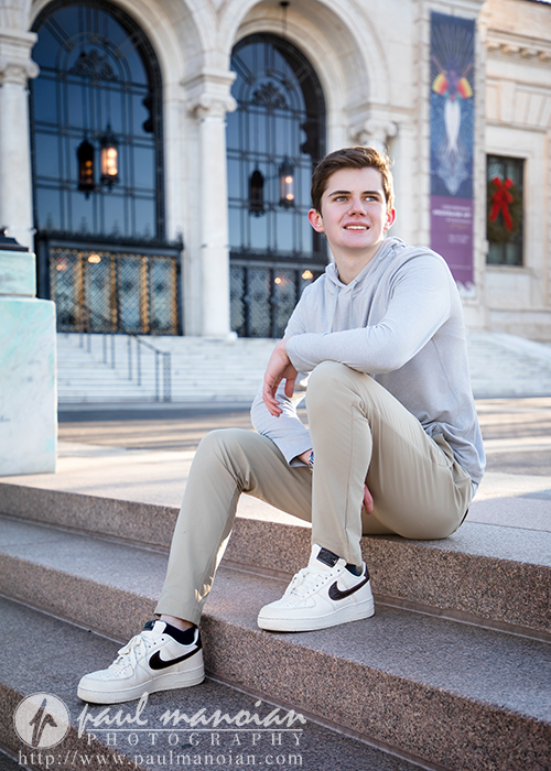 A young man in a light hoodie and khaki pants sits on stone steps outside a large, ornate building with arched doorways and banners, smiling to the side—capturing a candid moment from his Oxford senior pictures session.