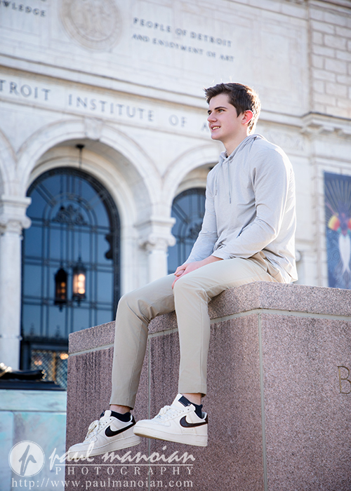 A young man in a light gray hoodie and khaki pants sits on a stone ledge outside the Detroit Institute of Arts, smiling and looking into the distance on a sunny day during his Oxford senior pictures session.