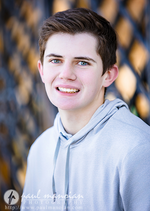 A teenage boy with short brown hair and blue eyes smiles in a light gray hoodie during his Oxford senior pictures session, standing outdoors before a blurred background with a subtle crisscross pattern.