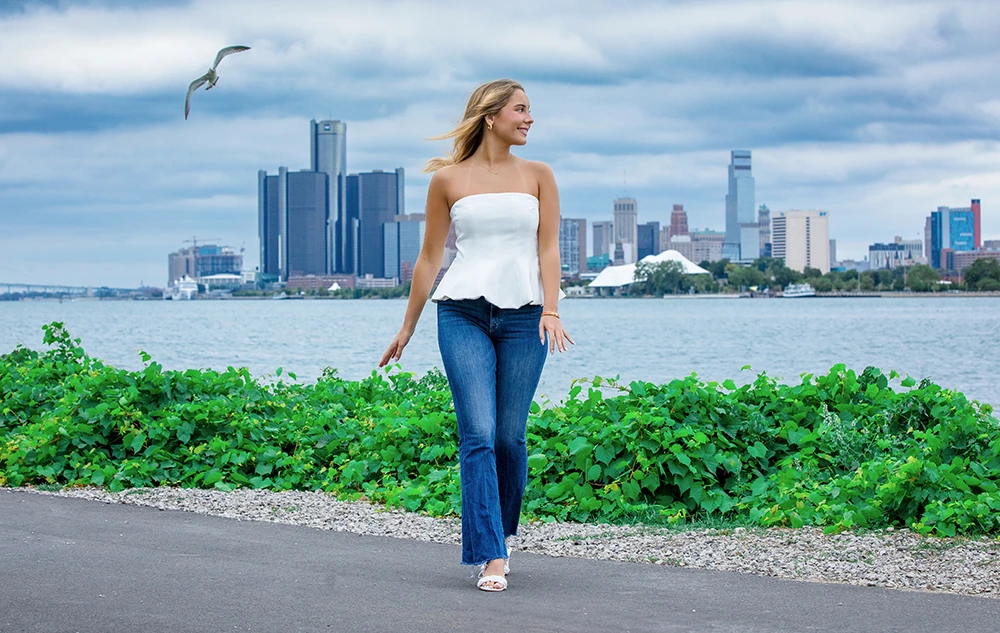 Senior Pictures Outfit Ideas for Girls in 2026: A high school senior girl in a white strapless top and blue jeans walks on a path by the water, with green plants and a city skyline in the background. A seagull flies overhead under a cloudy sky.