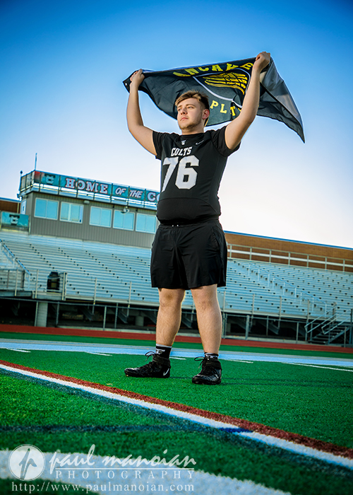 A football player in a black Colts uniform with number 76 holds a team flag above his head on a stadium field, capturing the spirit of a Troy senior pictures session under the clear blue sky with bleachers and a scoreboard behind him.